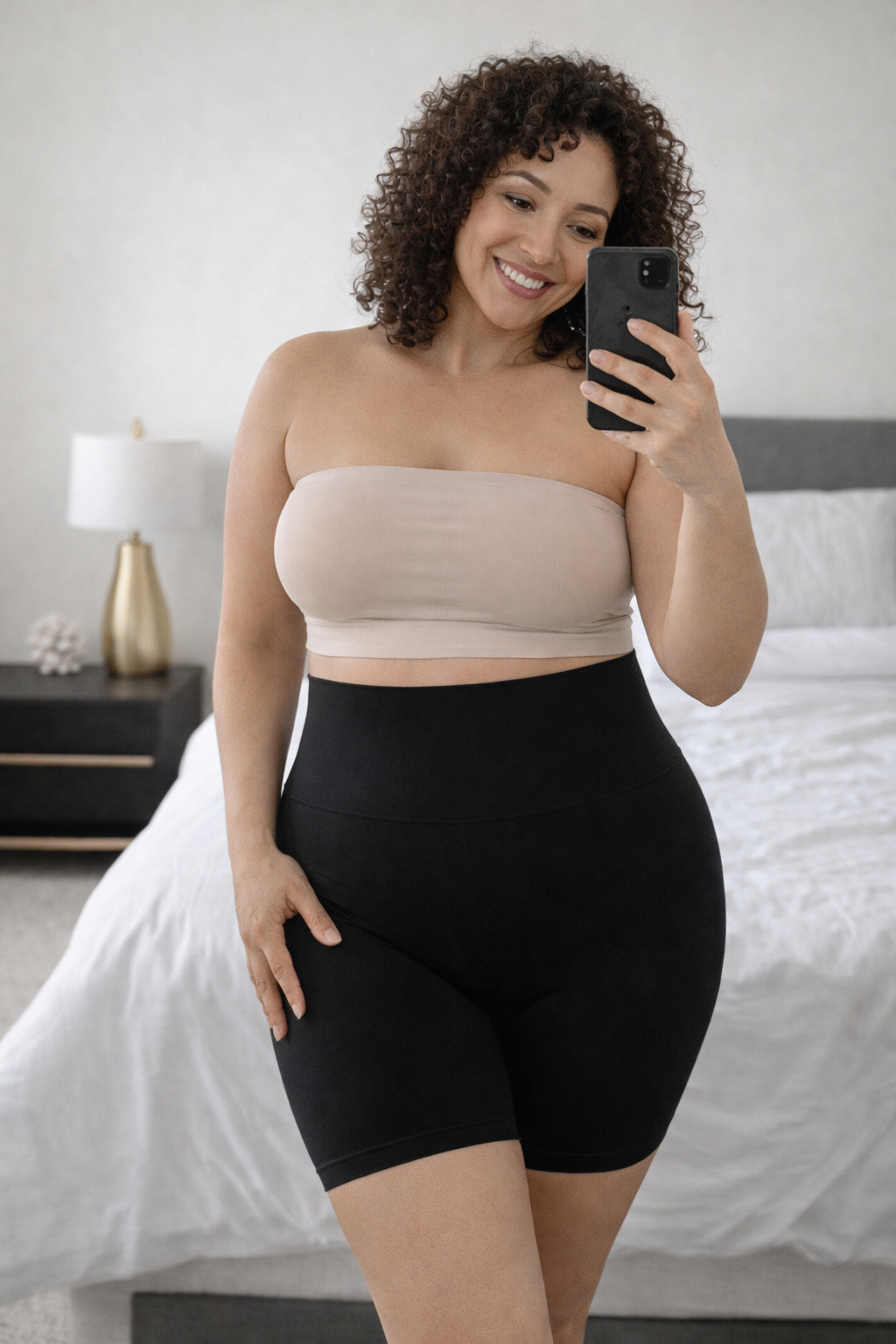 Woman taking a mirror selfie wearing Vellomi black PostureLift shaping shorts and a nude bandeau top in a bedroom setting.